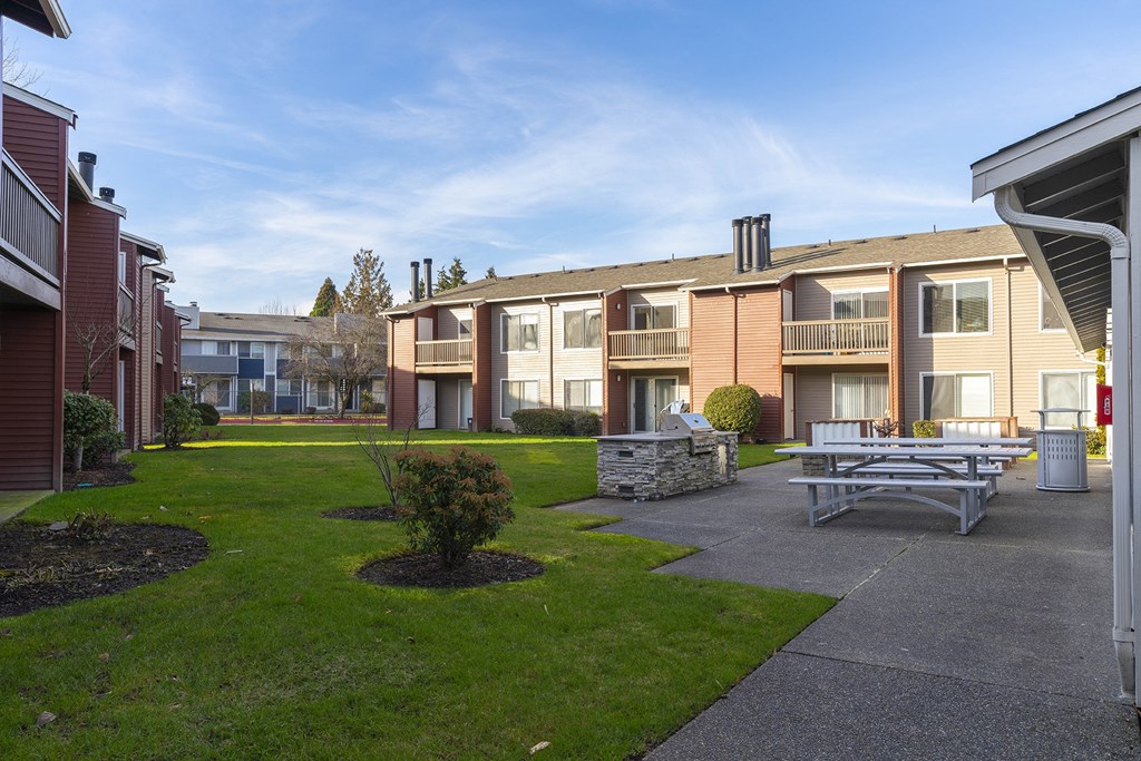A courtyard with picnic tables, a BBQ, and green grass with apartments in the background. at Pointe East, Fife, WA