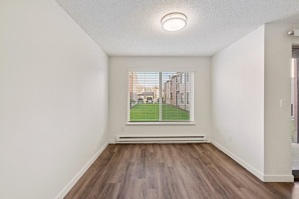 A dining room with white walls, wood flooring, overhead lighting, and an oversized window with a view of a bright green lawn. at Pointe East, Fife, WA, 98424