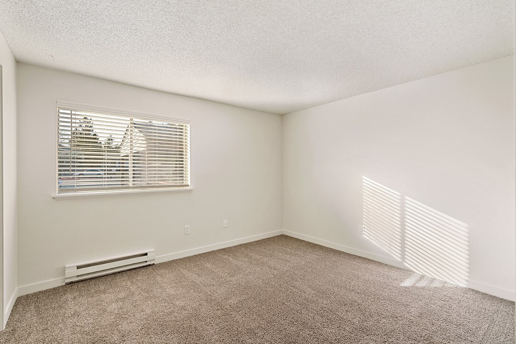 A large bedroom with white walls, taupe carpets, and a sunny window with white blinds. at Pointe East, Fife, Washington