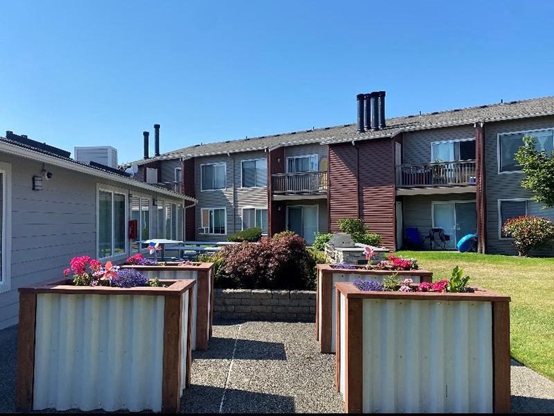 A row of four wooden planters with flowers in front of Pointe East Apartments with patios and balconies. at Pointe East, Fife, 98424