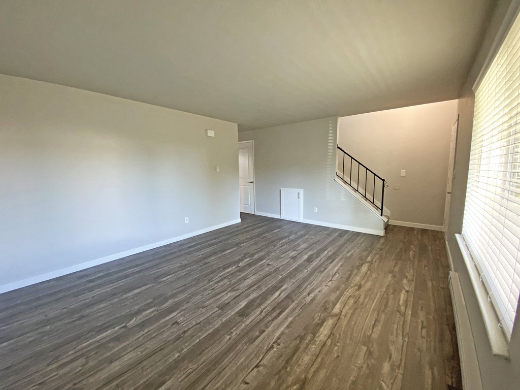 Large living room with faux wood flooring, a large window on the right wall, and an upstairs stairwell on the back wall.at Quilceda Gardens, Marysville, Washington