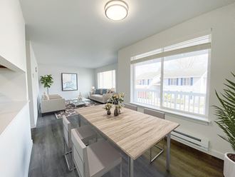 A dining room with white walls and a large sunny window that looks into the living room. Light-filled apartment staged with dining set and living room furniture.at The Trail, Snohomish, WA