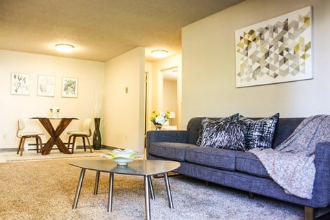 Carpeted living room with hallway that leads to bedrooms. Staged with grey couch, coffee table and art.at The Lakes Apartments, Washington, 98837