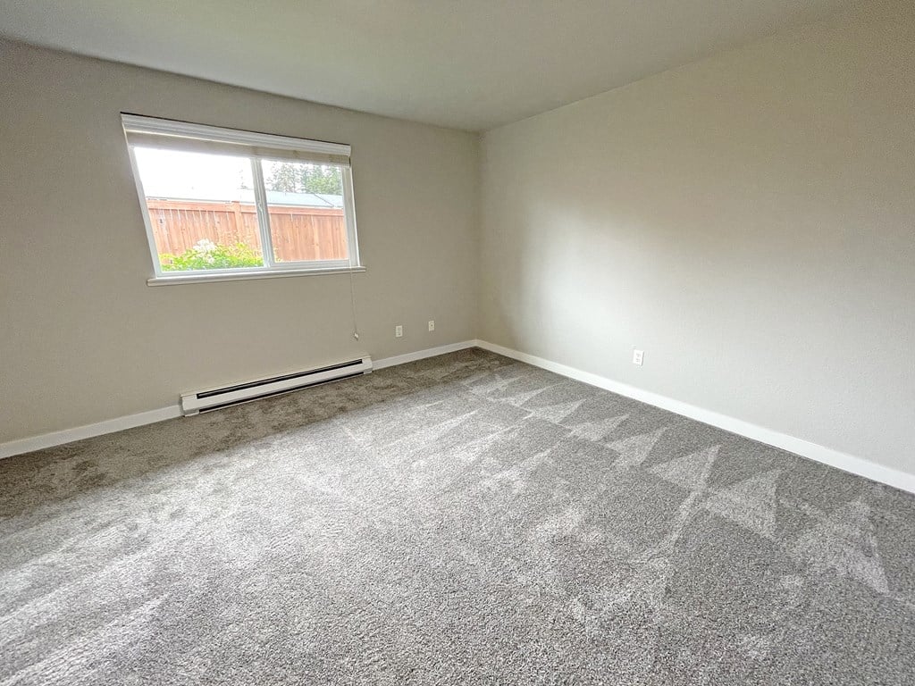 Light-filled bedroom corner, gray carpet, a large window on the left wall with heater below.at Quilceda Gardens, Marysville, 98270
