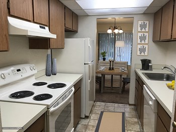 Galley kitchen with brown cabinets and white appliances looking into dining area. - Photo Gallery 5