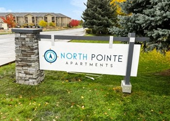 Outside entrance with a blue and white North Pointe sign surrounded by grass, trees, and a road to apartments behind. - Photo Gallery 16