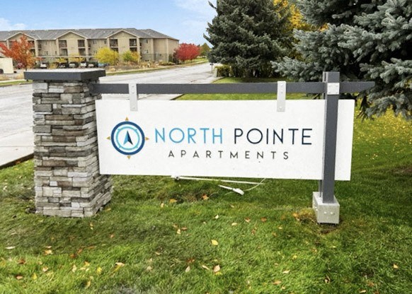 Outside entrance with a blue and white North Pointe sign surrounded by grass, trees, and a road to apartments behind.at North Pointe, Post Falls, ID