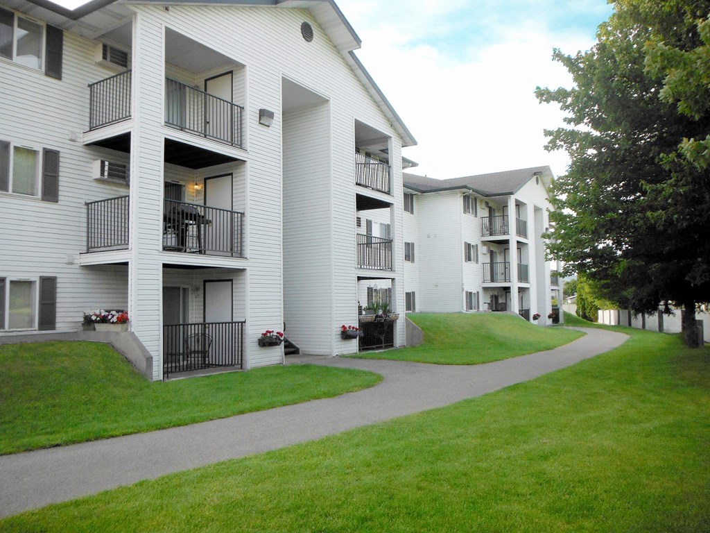 North Pointe Apartments exterior with covered patios and balconies surrounded by fenced green grass, a paved path, and trees.at North Pointe, Post Falls