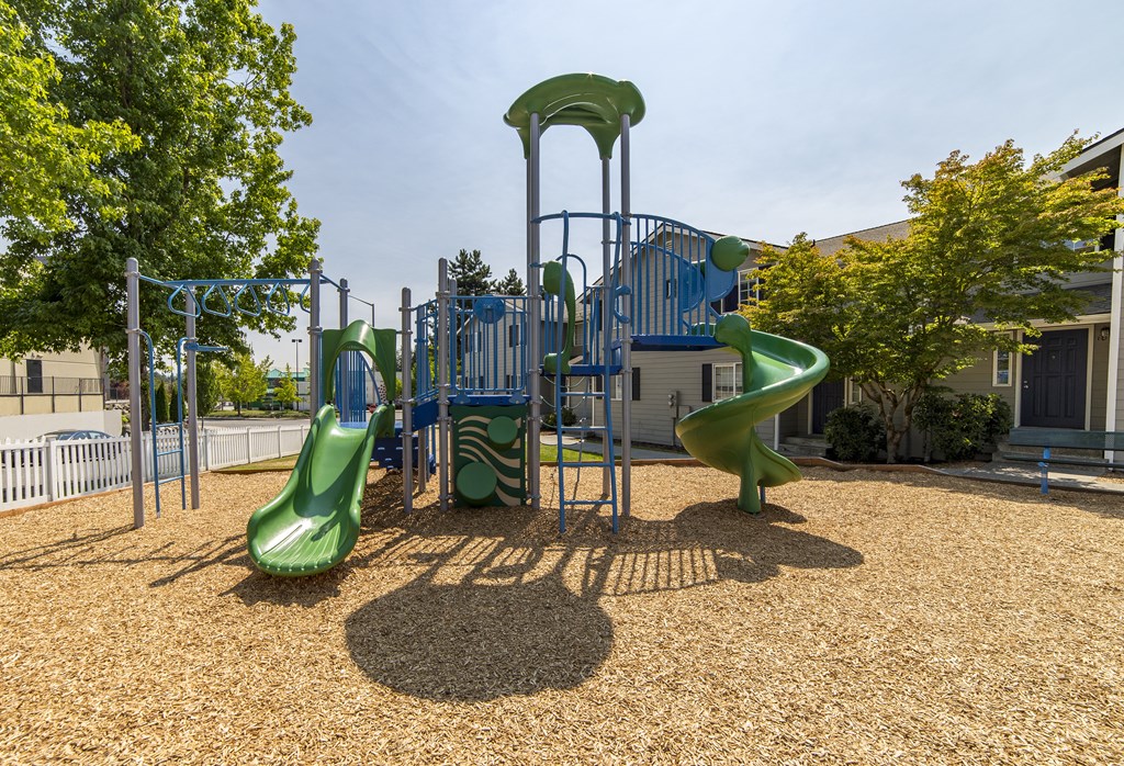 Green and blue playground surfaced with wood chips and enclosed by a white picket fence and green trees at Arterra Apartments, Kent, WA