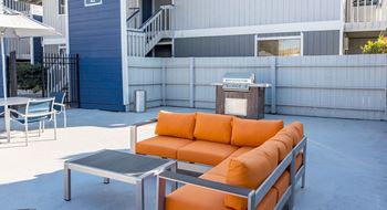Outdoor resident lounge with organge patio couch, a coffee table, umbrella covered seating and a BBQ.at The Lakes Apartments, Moses Lake, WA