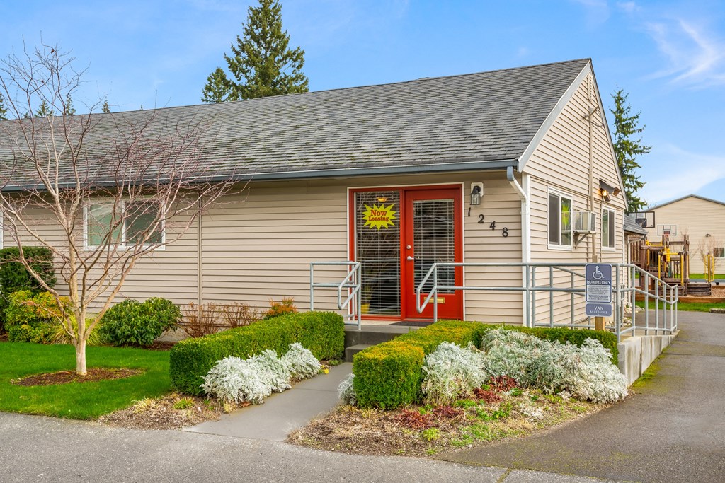 Outside of Leasing Office, red doors, a 'Now Leasing' sign, a wheelchair ramp,  landscaping, basketball court behind. at Quilceda Gardens, Washington