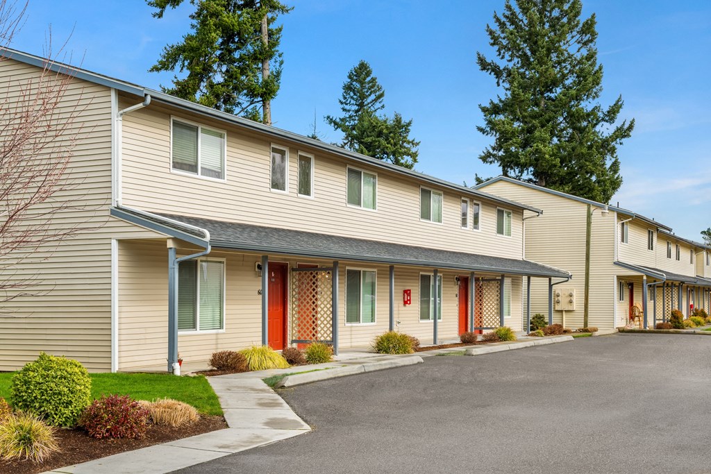 Exterior apartments lined with a paved walkway, maintained landscaping, trees in the distance, and parking spaces in front.at Quilceda Gardens, Marysville, WA 98270
