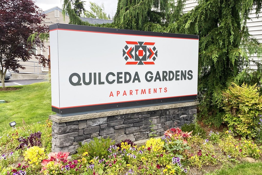 Community entrance with red and gray 'Quilceda Gardens' sign surrounded by flowers, grass, and trees.at Quilceda Gardens, Marysville