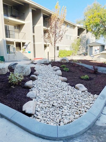 Outside of community with well-maintained landscaping, decorative rocks path, plants, and a paved path to apartments behind. - Photo Gallery 17