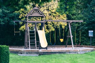 Playground with a yellow slide, three swings, a trapeze, and a latter. Surrounded by grass, trees, and landscaping.at Sitka Heights, Fife, 98424