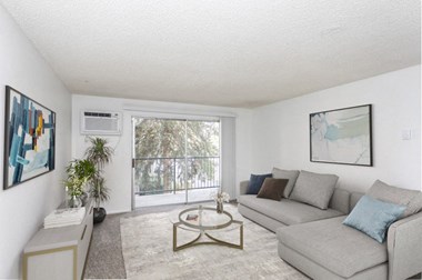 a spacious living room white walls, taupe carpet, and huge double sliding glass doors to private balcony. Staged with a gray couch, a coffee table, console table and wall art.