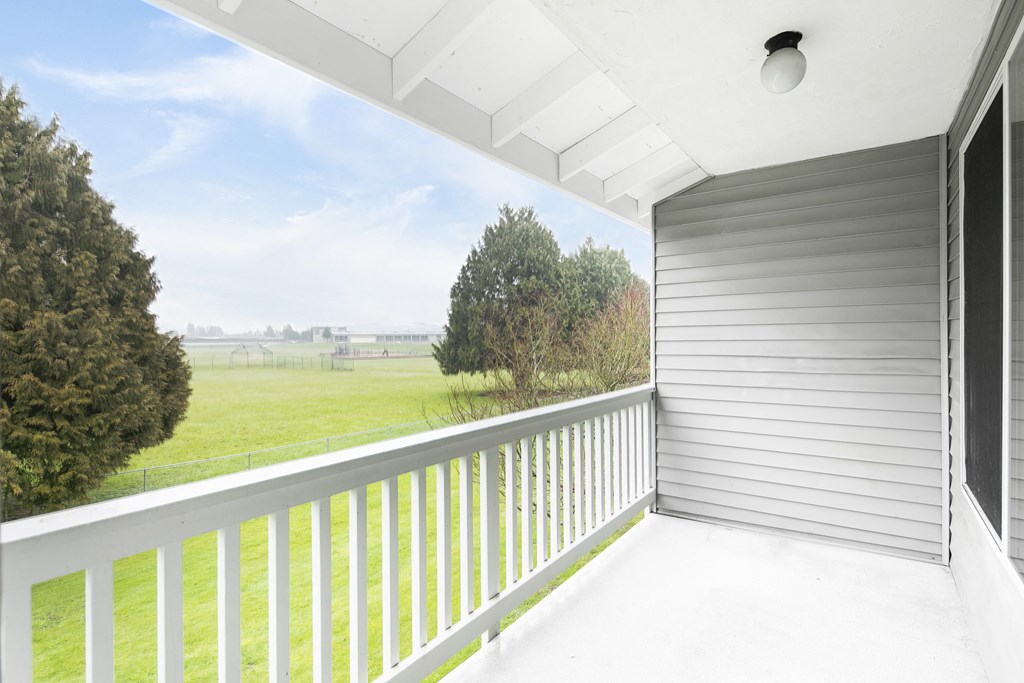 A private, covered apartment balcony with a white railing looks out to grass fields and trees.at The Trail, Snohomish, 98290