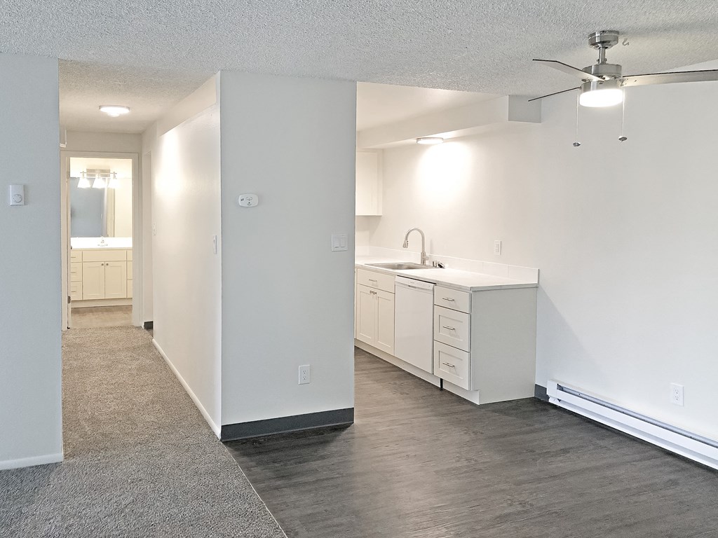 Apartment with a hallway on the left and kitchen on the right with faux wood flooring, white cabinets and appliances and a ceiling fan.