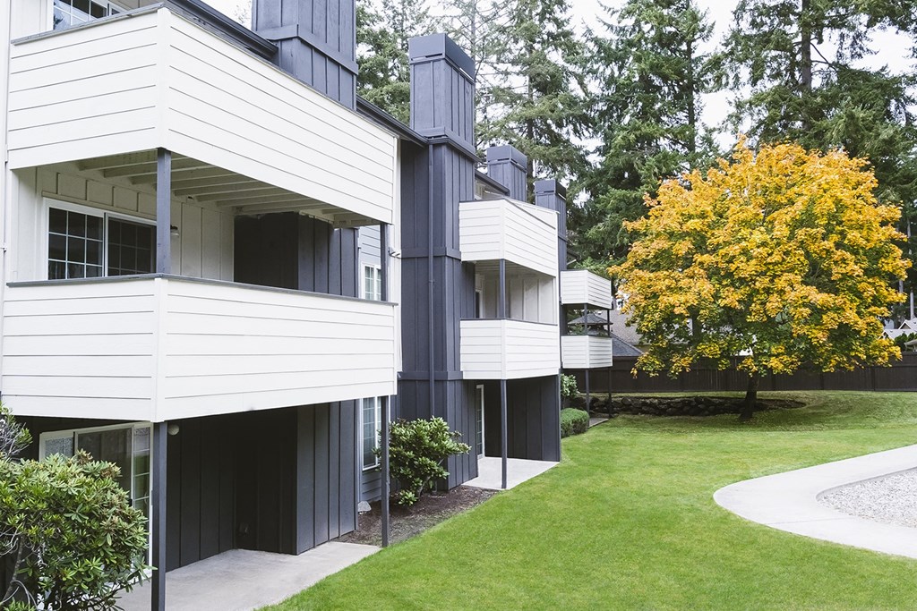 Apartments with patios and balconies lined by manicured lawn, landscaping, a large tree, and a curved paved pathway at University Glen, University Place, 98466
