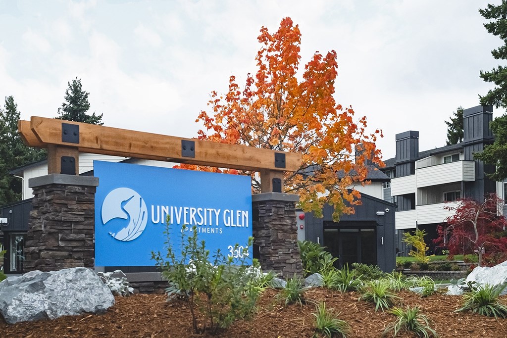 Community entrance with blue 'University Glen' sign surrounded by green plants, bushes, large rocks, and trees at University Glen, University Place, WA
