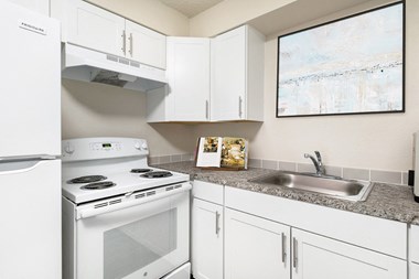 Corner of a kitchen with custom white cabinets, spacious countertops, a stainless steel sink, oven, stovetop and hood range, and full-size fridge.