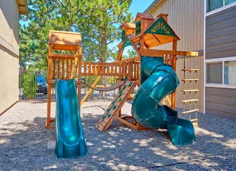 A playground with two slides, a swing set, and a small rock climbing wall surrounded by pea gravel and trees in the background.at Wildflower, Washington