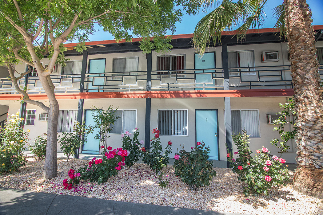 Exterior Building lined with rock garden, pink and red flowers and palm trees at Woodland Village Apartments , Woodland, CA 95695
