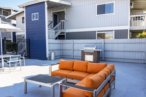 Outdoor lounge with umbrella-covered tables, gas BBQ, and an oversized orange patio couch and coffee table.at The Lakes Apartments, Moses Lake, WA