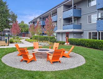 Exterior view of the building and community area. Outdoor area w/ brick fireplace with 6 orange lawn chairs around it. Fire pit space is surrounded by green grass with a path leading up to it.