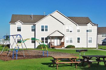 A playground with a swing set and picnic tables is in front of a white building.