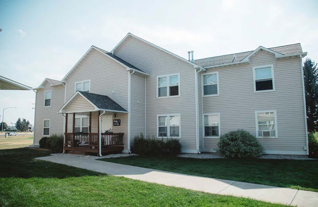 Apartment building exterior with a front porch, decorative gravel, green grass, and bushes.