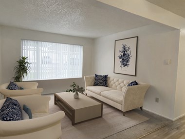 Carpeted living room with oversized window, white panel track blinds, staged with couch, coffee table, 2 chairs, and wall art.