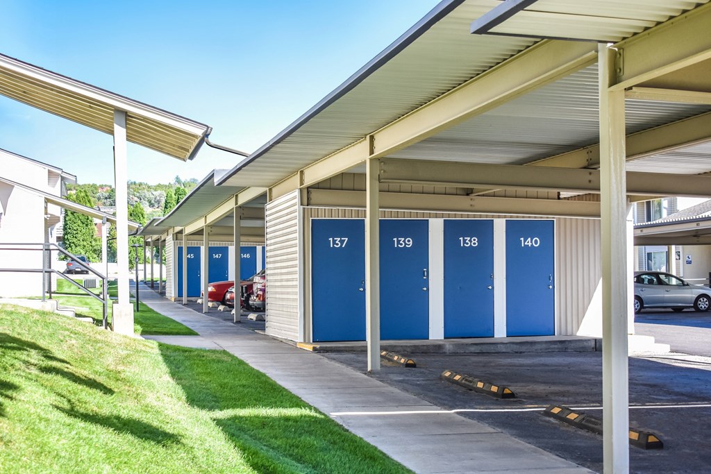 Covered parking and storage units with blue doors next to sidewalk lined with green grass in Pocatello, ID