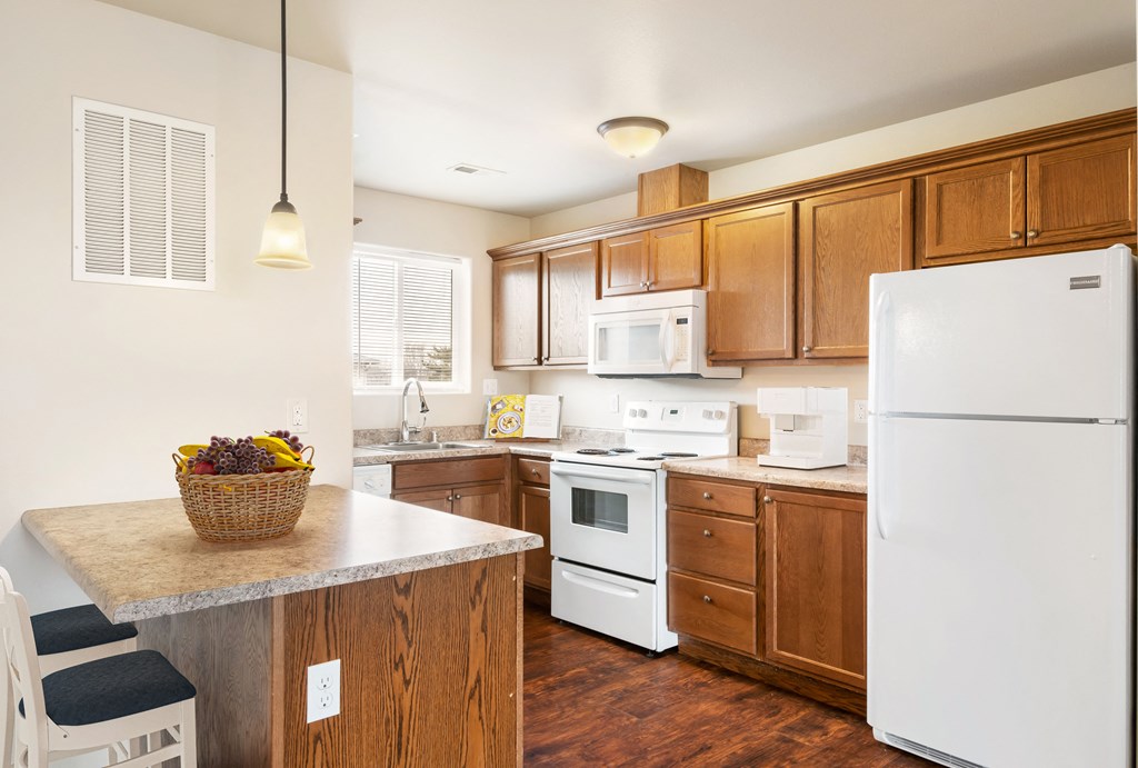 L-shaped kitchen with top and bottom cabinet storage, white appliances, and a breakfast bar staged with bar stools. at Pine Tree Park, Kennewick