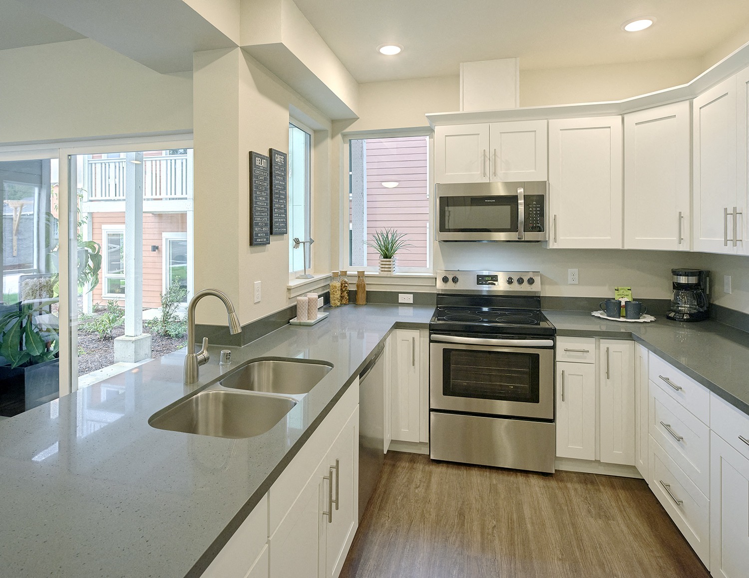 Light-filled u-shaped kitchen with two large windows, white cabinets, double bowl sink, oven, stovetop, and microwave.