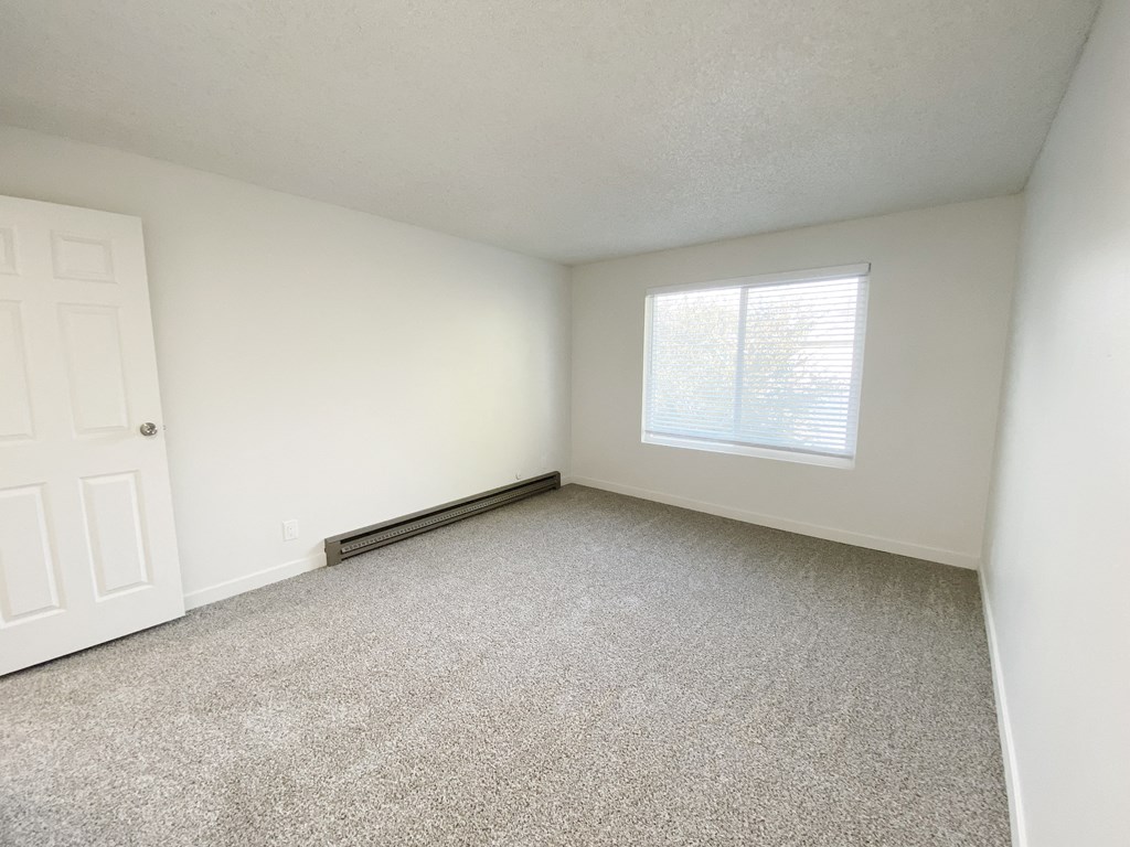 Bedroom with gray carpets, a heater on the left wall, and a large window with blinds on the back wall.at Laurel Park, Twin Falls