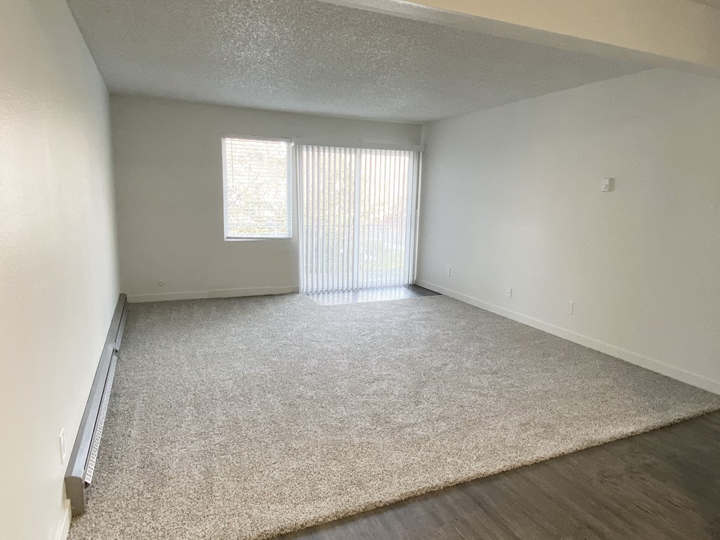 Open living room with gray carpets, a window, and sliding glass door to balcony with nature views on the back wall.at Laurel Park, Twin Falls, Idaho