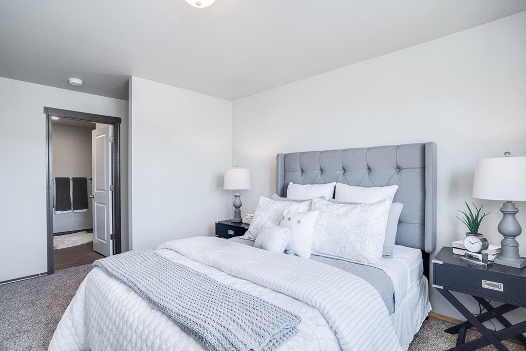 Large carpeted bedroom with full ensuite bathroom on the left. Staged with a queen bed, side tables, and lamps.at North Pointe, Idaho