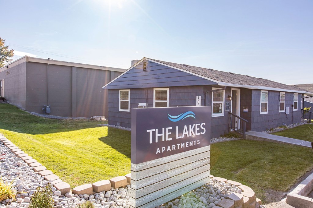 Outside of community with a blue 'The Lakes Apartment' sign surrounded by decorative rocks, green grass, and leasing office behind.at The Lakes Apartments, Moses Lake, WA
