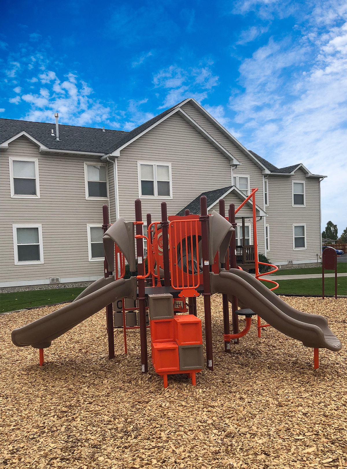 Orange and brown playground surfaced with woodchips and surrounded by green grass and paved pathways.