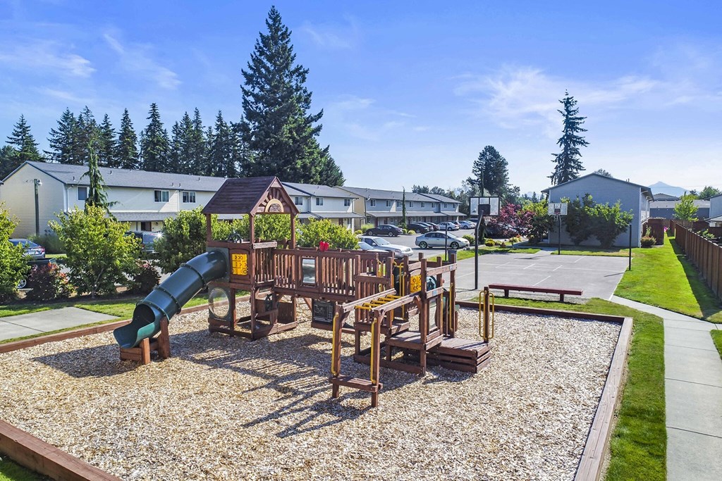 Large outdoor community playground with green slide, surrounded by manicured lawn with full basketball court behind.at Quilceda Gardens, Marysville, WA