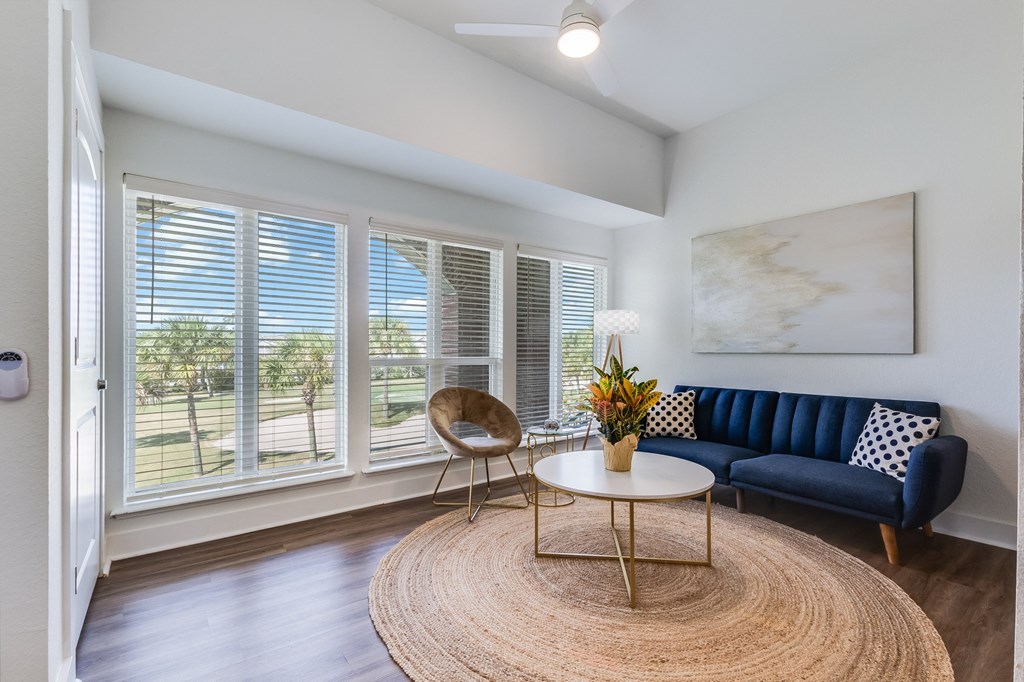 A living room with a blue couch, a round table, and a large window with blinds.
