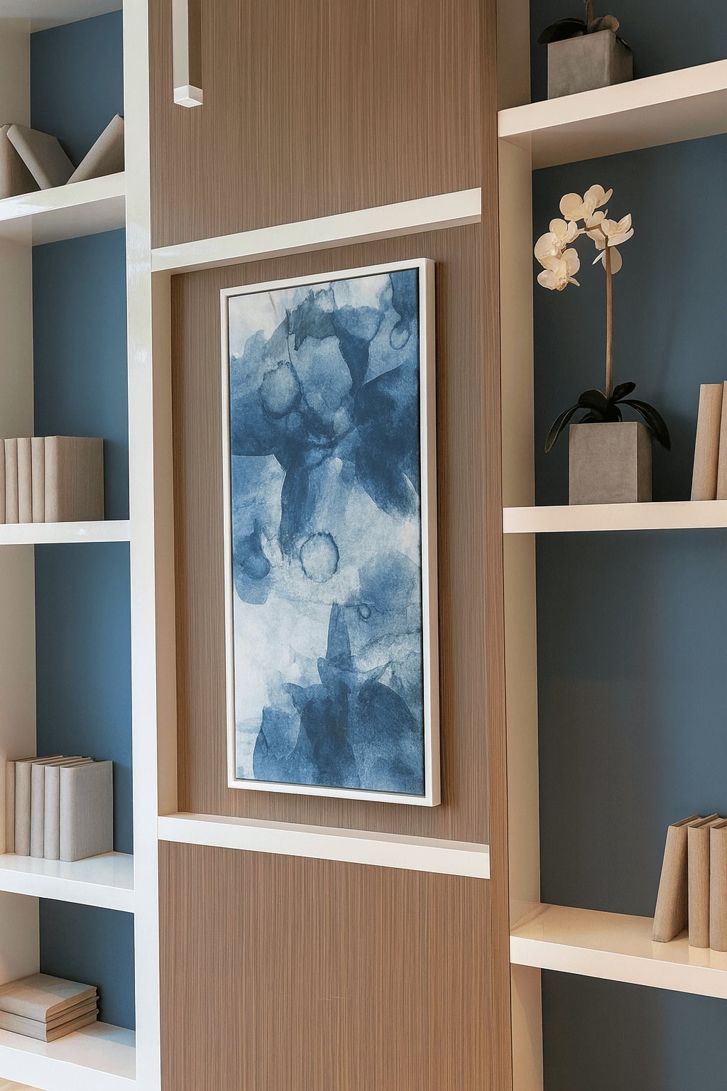 A framed artwork hangs on a wall above a shelf with a flower and books.