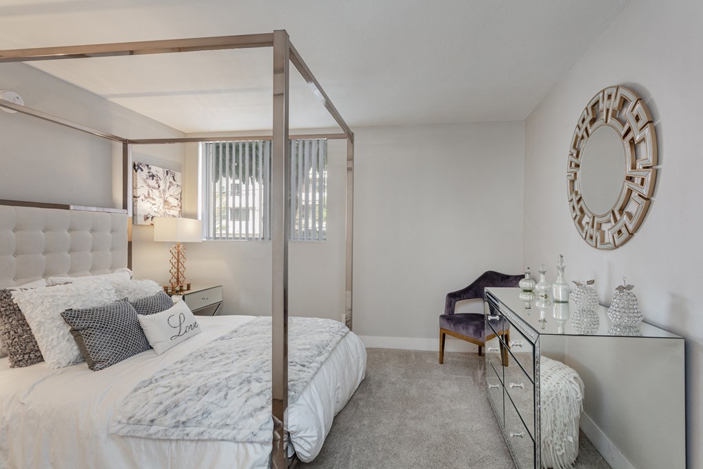 Hollywood, CA, Apartments with In-Unit Washer and Dryer - Candela - Bedroom with Carpet Flooring, Large Four-Poster Bed with Pillows, Nightstand, Lamp, Wall Art, Round Wall Mirror, Armchair, Mirrored Dresser, Home Decor, and a Window with Horizontal Blinds.