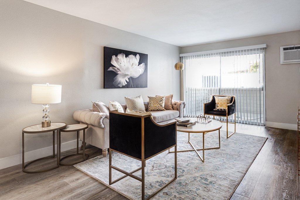 Dog-Friendly Apartments in Hollywood, CA - Candela - Living Room with Luxury Vinyl Plank Flooring, Couch, Accented Black Armchairs, Round Coffee Table, Large Rug, Round Nested Side Tables, Lamp, Large Wall Art, Air Conditioning Unit, and a Large Sliding Door with Blinds Leading to the Balcony.