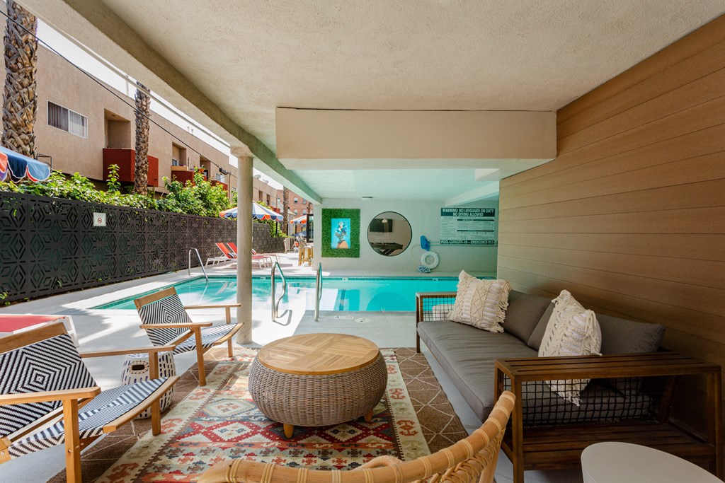 Two-Bedroom Apartments in Hollywood, CA - Candela - A Covered Outdoor Lounge Area Furnished with Patterned Lounge Chairs, Long Wooden Cushion Couch with Pillows, Round Wood-Top Woven Coffee Table, Large Rug, and a View of the Rectangular Swimming Pool.