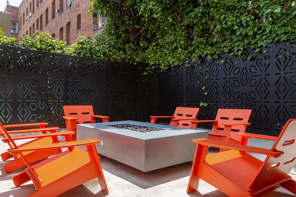 Pet-Friendly Apartments in Hollywood, CA - Candela - Modern Outdoor Patio Featuring a Rectangular Steel Fire Pit at the Center of 6 Orange Wooden Lounge Chairs, Surrounded by Black Decorative High Fence, with a View of the Greens and Apartments.