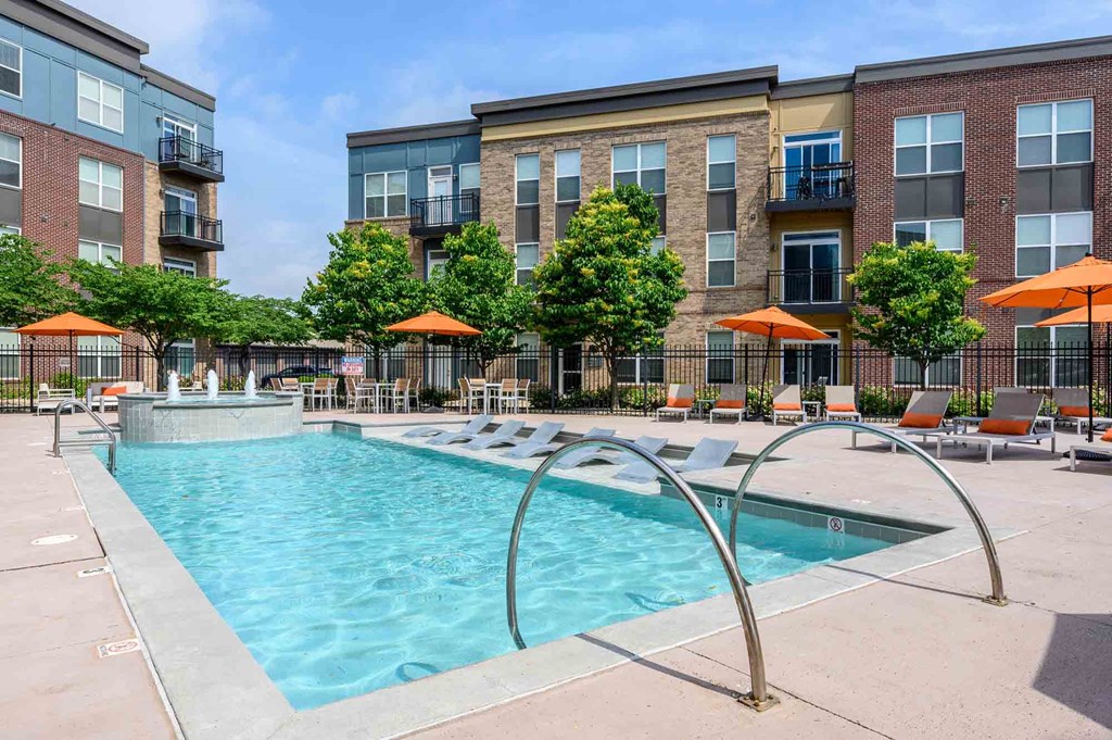 A swimming pool surrounded by orange umbrellas and lounge chairs in front of apartment buildings.