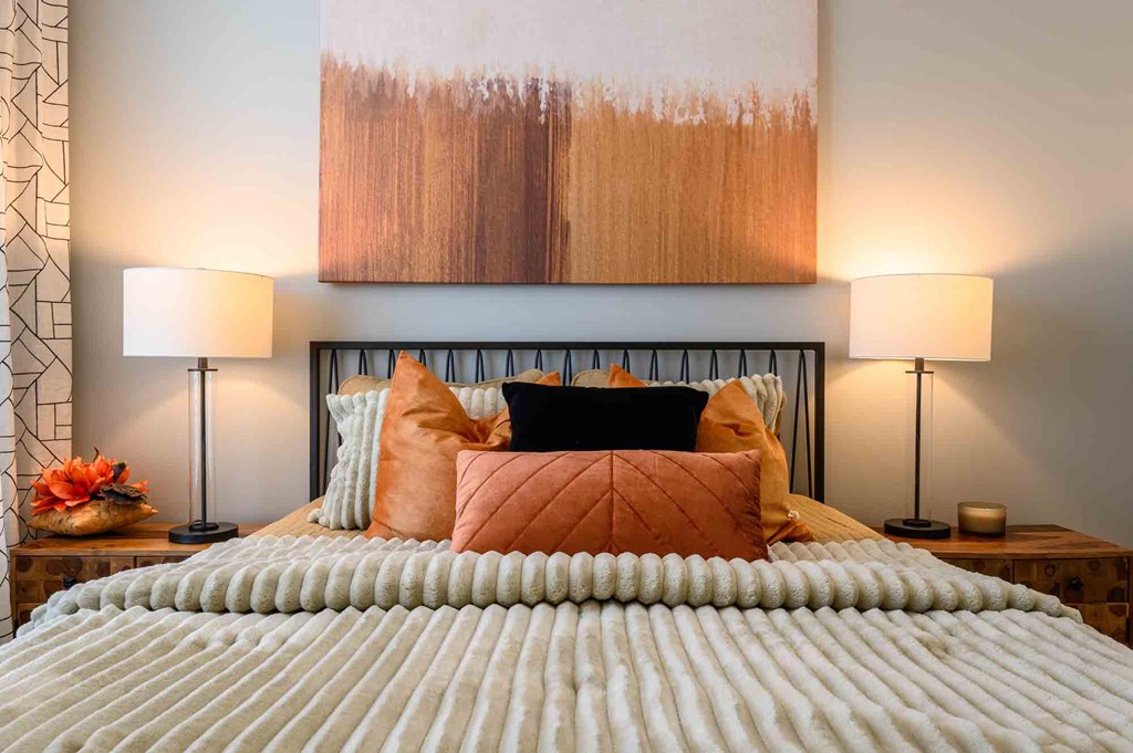 A bed with a white comforter and orange pillows is in the foreground with two lamps on each side.