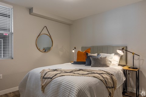 1-Bedroom Apartments in Vancouver, WA - 134th Street Lofts - Bedroom with Cream Walls, Bed, Nightstands with Lamps, Round Wall-Mounted Mirror, and Window.